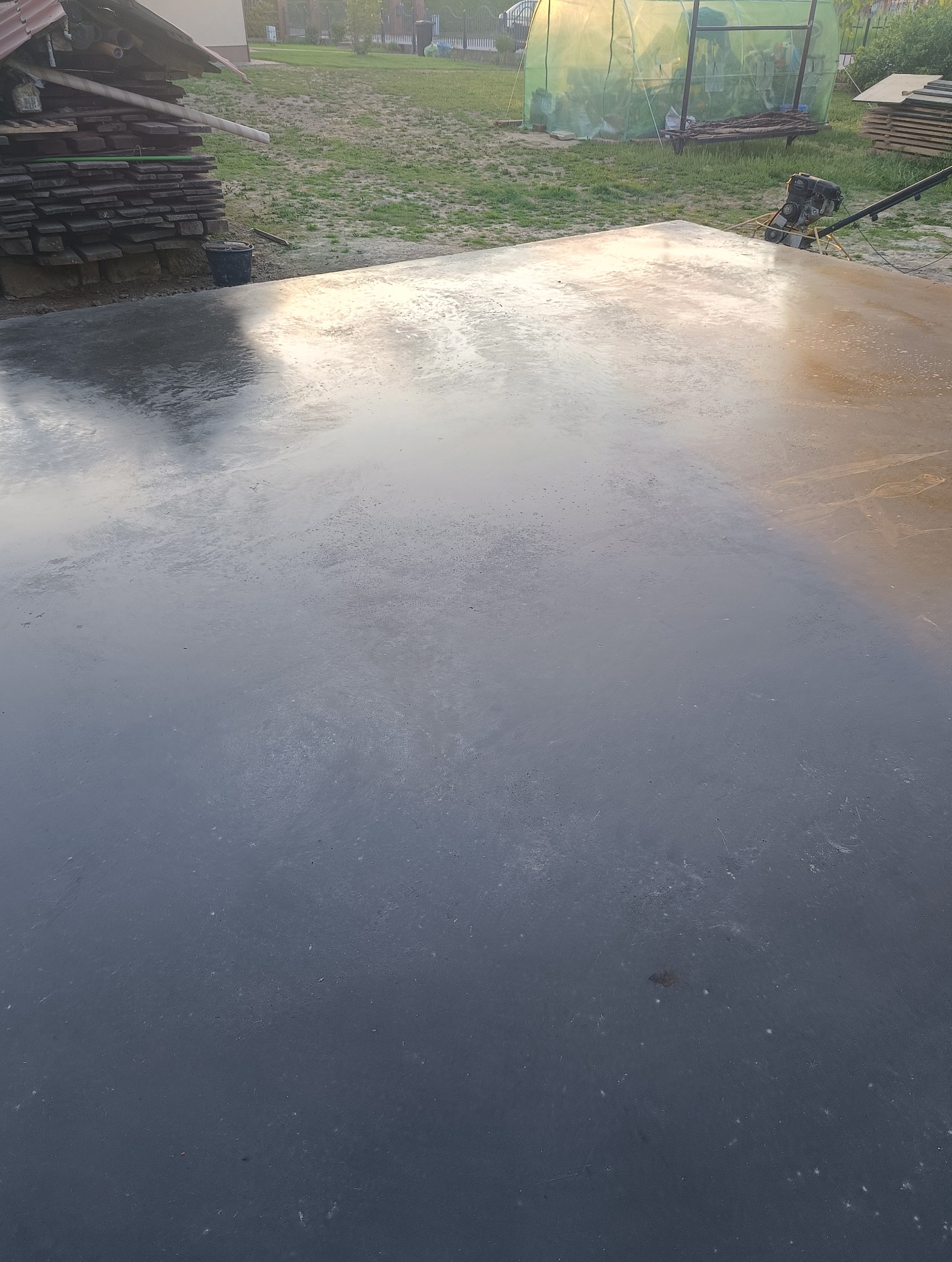 Świeżo wylana, mokra posadzka betonowa w trakcie zacierania mechanicznego zacieraczką, widoczna sterta desek i szklarnia w tle.