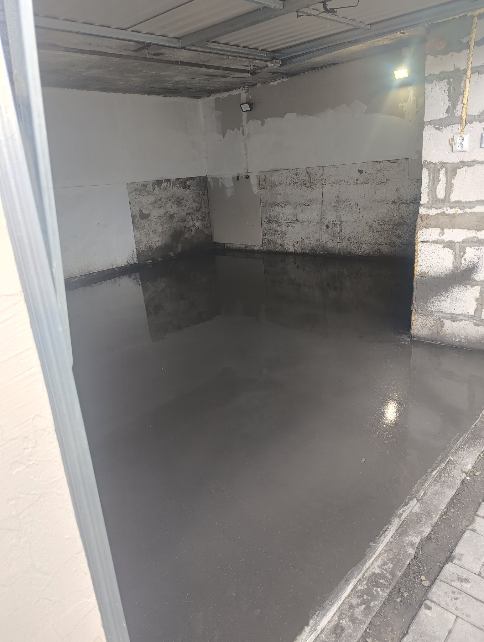 Świeżo wylana, gładka, szara posadzka w surowym pomieszczeniu z niedokończonymi ścianami z betonu komórkowego i częściowo otynkowanymi fragmentami, widoczne gniazdka elektryczne i lampa sufitowa.