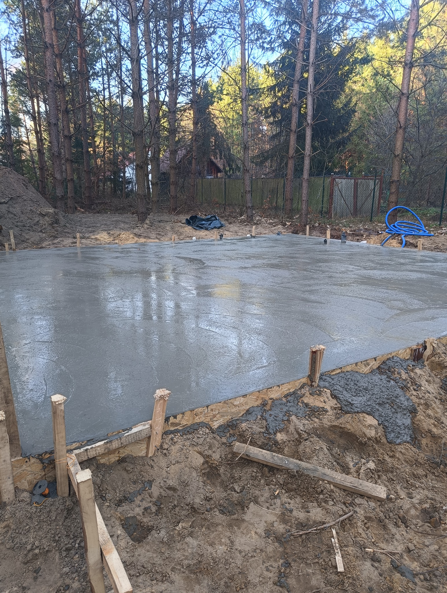 Świeżo wylana, gładka płyta fundamentowa z drewnianym szalunkiem i wystającymi kołkami, otoczona ziemią i drzewami w tle.