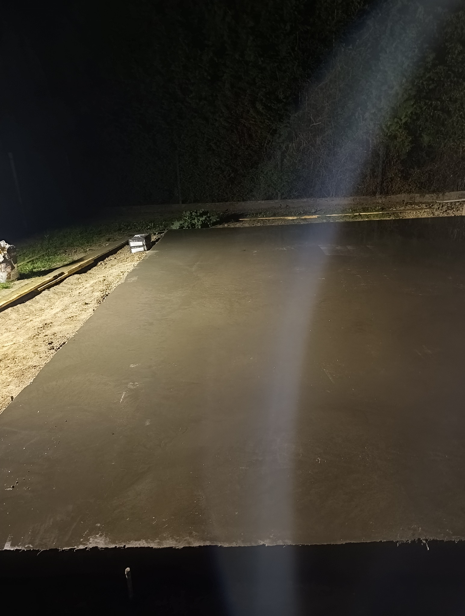 Świeżo wylana, gładka posadzka betonowa, oświetlona punktowo nocą, z widocznymi śladami prac wykończeniowych na obrzeżach i deskami szalunkowymi w tle.