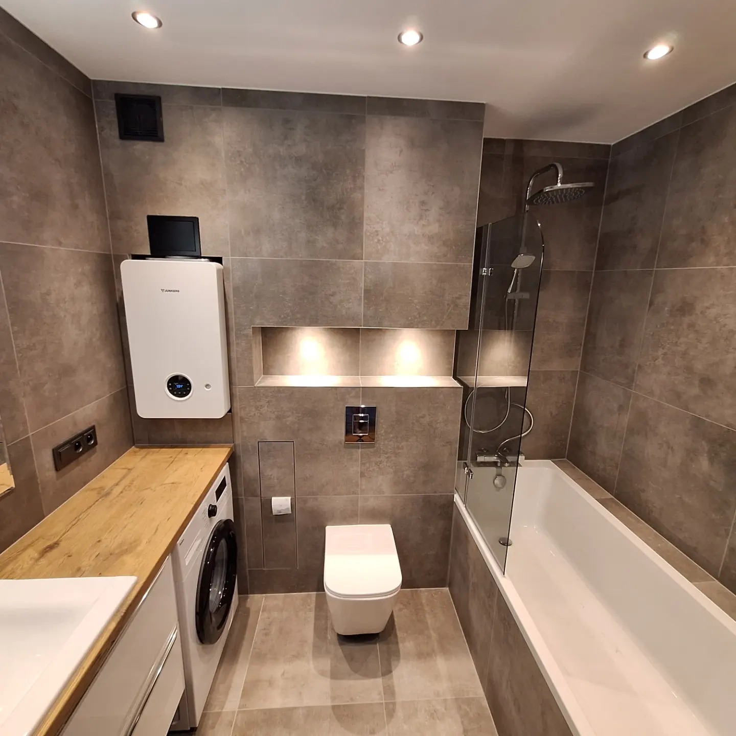 Nowoczesna łazienka z szarymi płytkami, białą wanną z parawanem, toaletą podwieszaną, pralką zabudowaną pod drewnianym blatem i piecem gazowym.