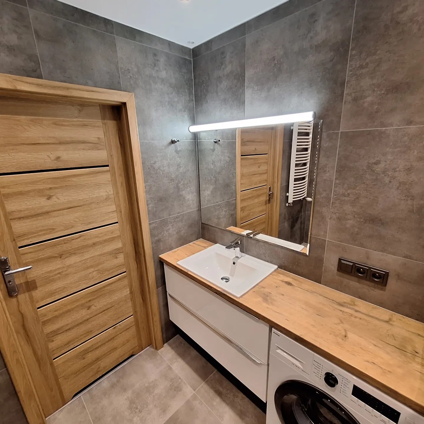 Nowoczesna łazienka z szarymi płytkami, drewnianym blatem, białą szafką pod umywalką i drewnianymi drzwiami z poziomymi wstawkami, widoczna pralka.