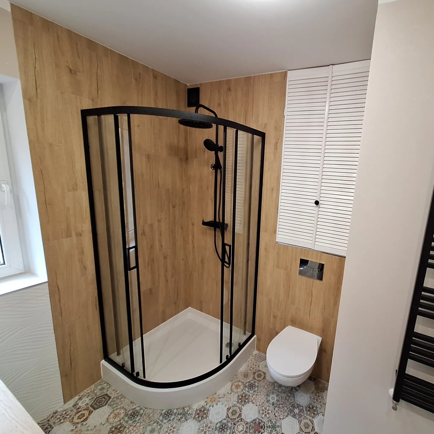 Wykończona łazienka z kabiną prysznicową z czarnymi profilami, toaletą podwieszaną i grzejnikiem drabinkowym w odcieniach drewna i bieli, na podłodze wzorzyste płytki heksagonalne.