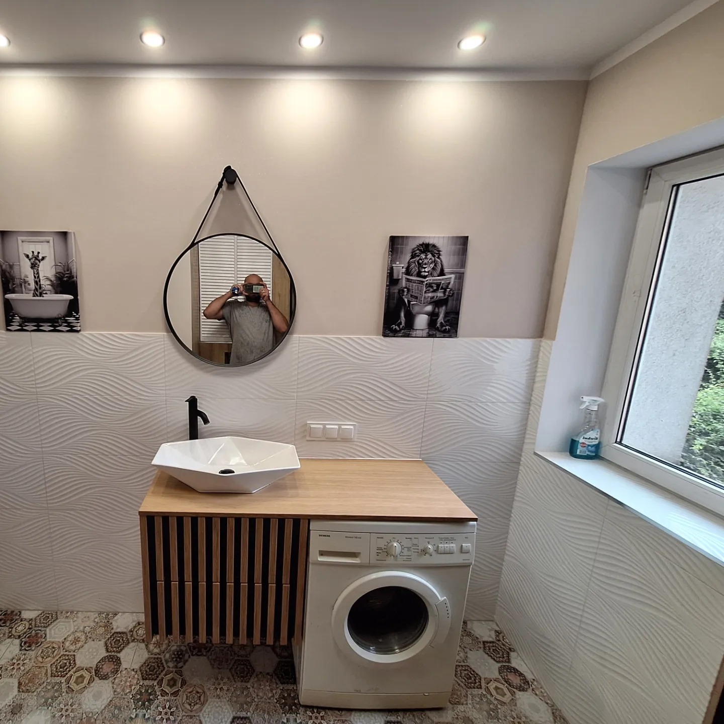 Nowoczesna łazienka z umywalką nablatową, szafką z lamelami, pralką i dekoracyjnymi obrazami na ścianie. W lustrze odbija się osoba robiąca zdjęcie.