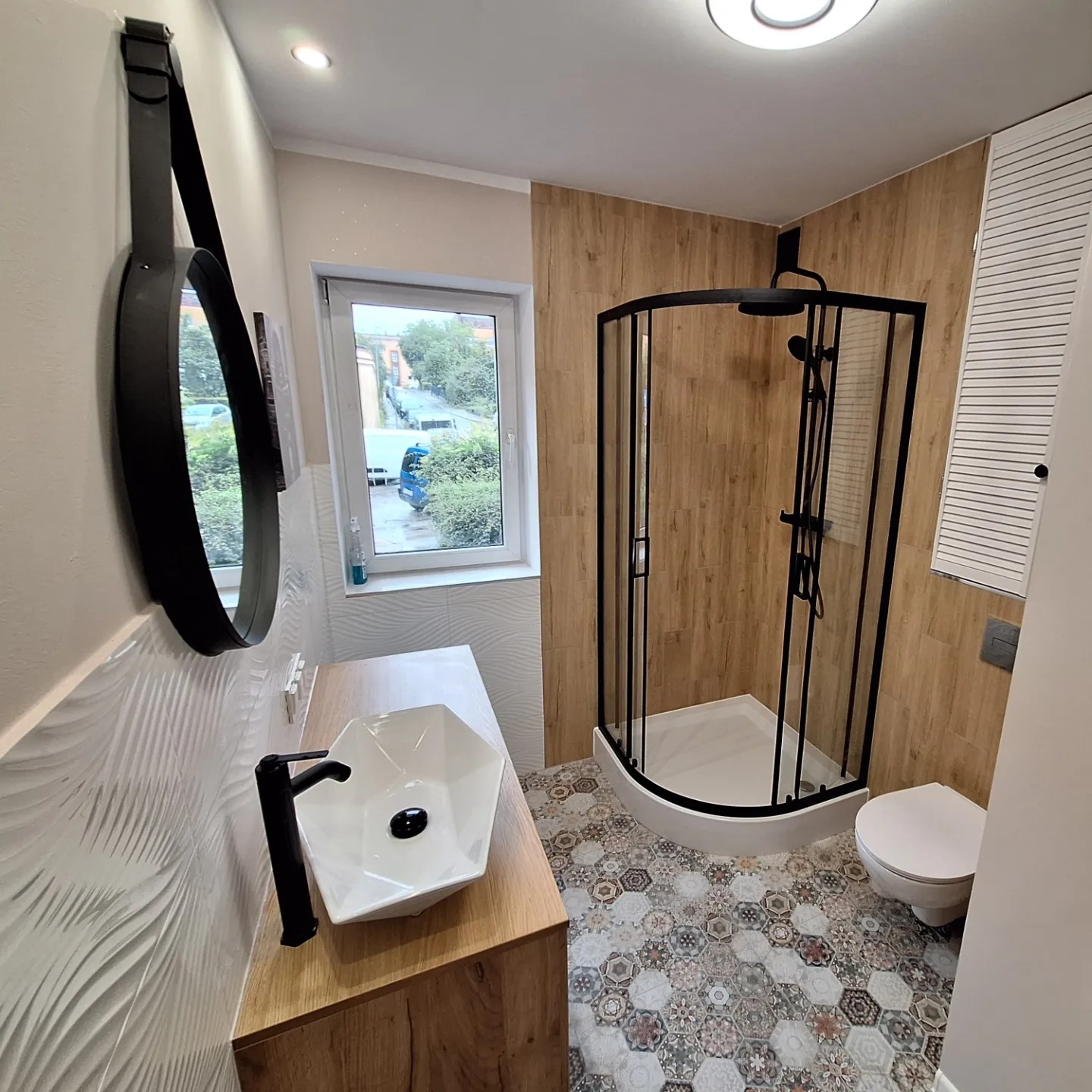Nowoczesna łazienka z kabiną prysznicową z czarnymi ramami, umywalką w kształcie diamentu na drewnianym blacie, okrągłym lustrem na pasku i wzorzystą podłogą.