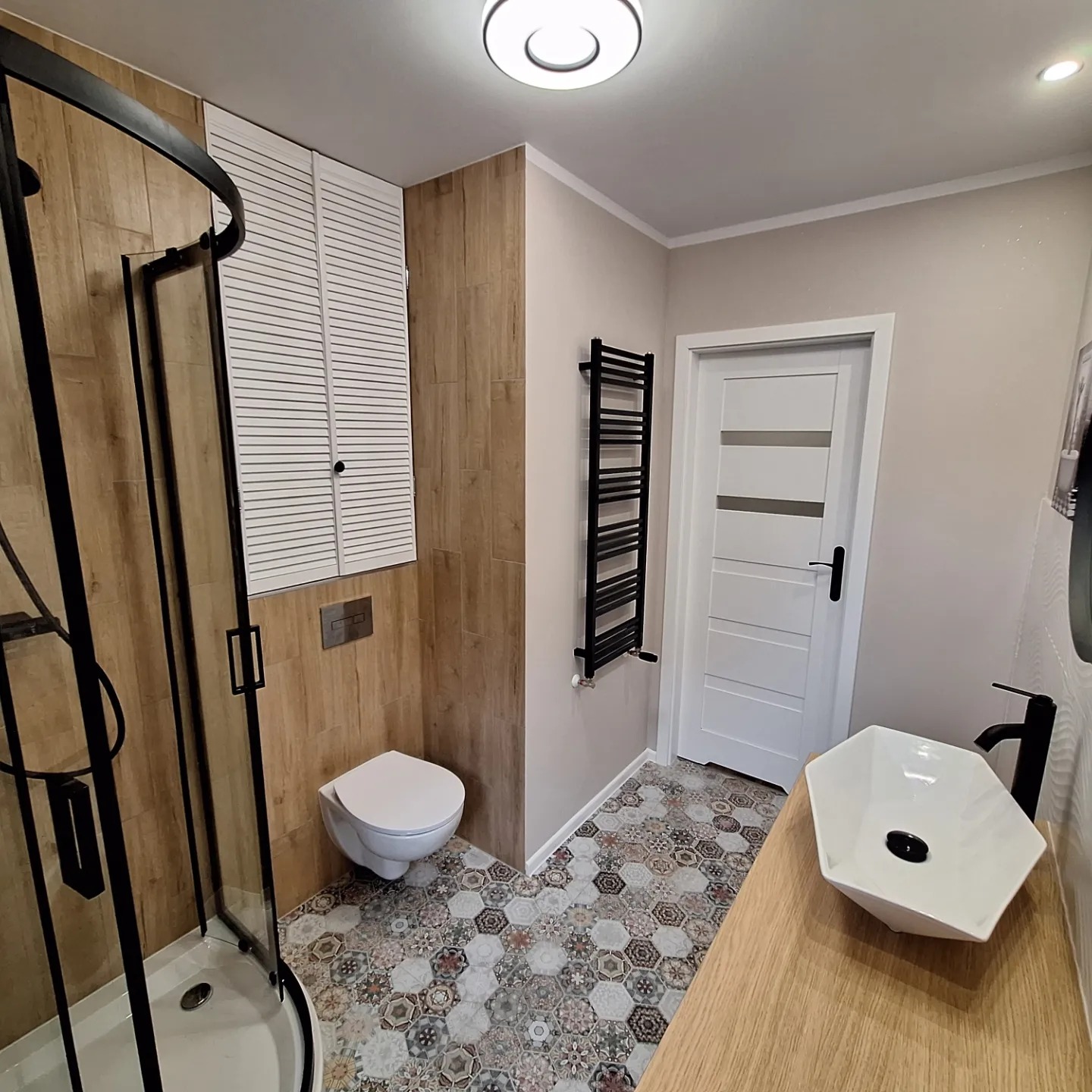 Nowoczesna łazienka z kabiną prysznicową z czarnymi ramami, wiszącą toaletą, grzejnikiem drabinkowym i umywalką nablatową na drewnianym blacie, wykończona płytkami heksagonalnymi.