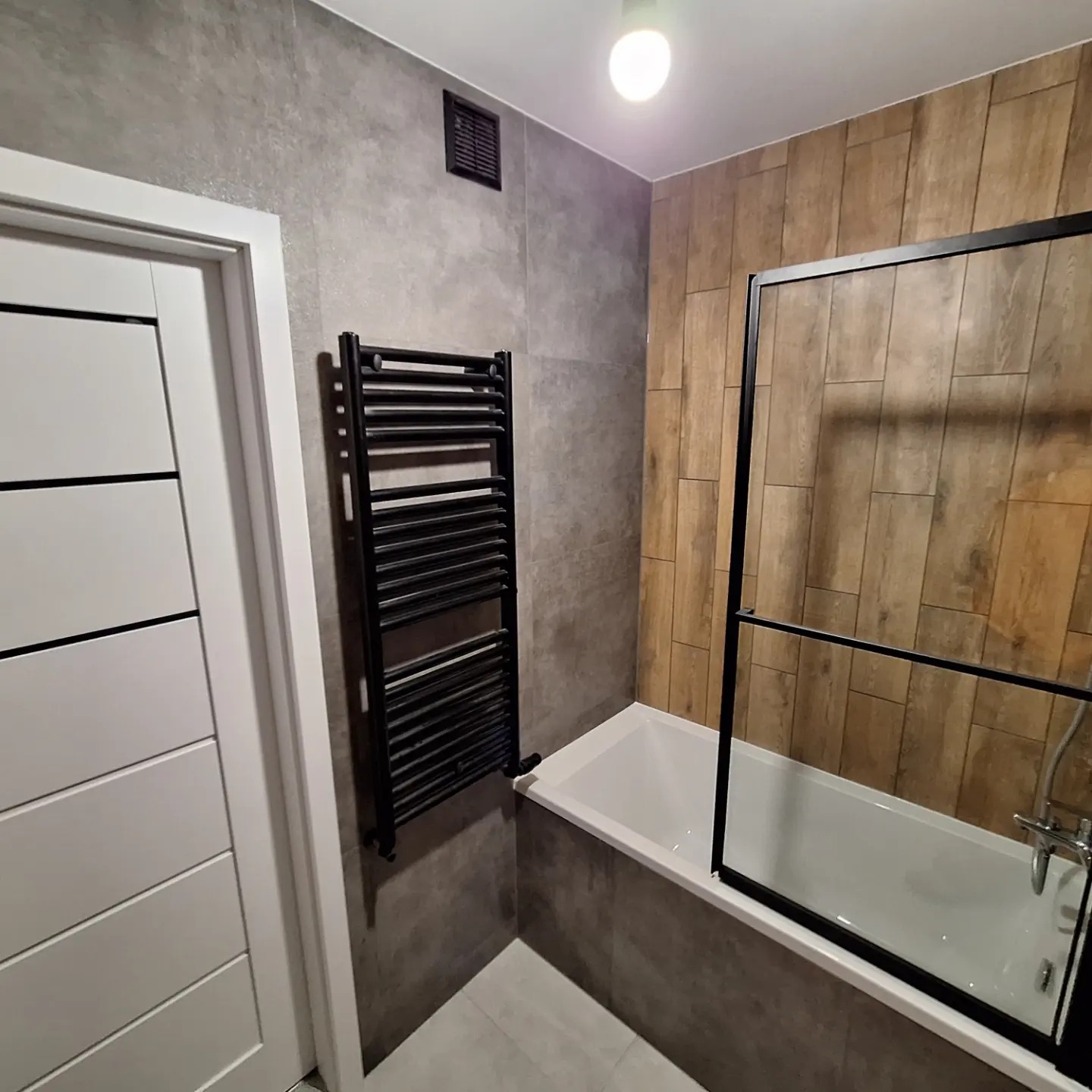 Nowoczesna łazienka z szarymi płytkami imitującymi beton, drewnopodobnymi panelami na ścianie, białą wanną z czarnym parawanem nawannowym oraz czarnym grzejnikiem drabinkowym.