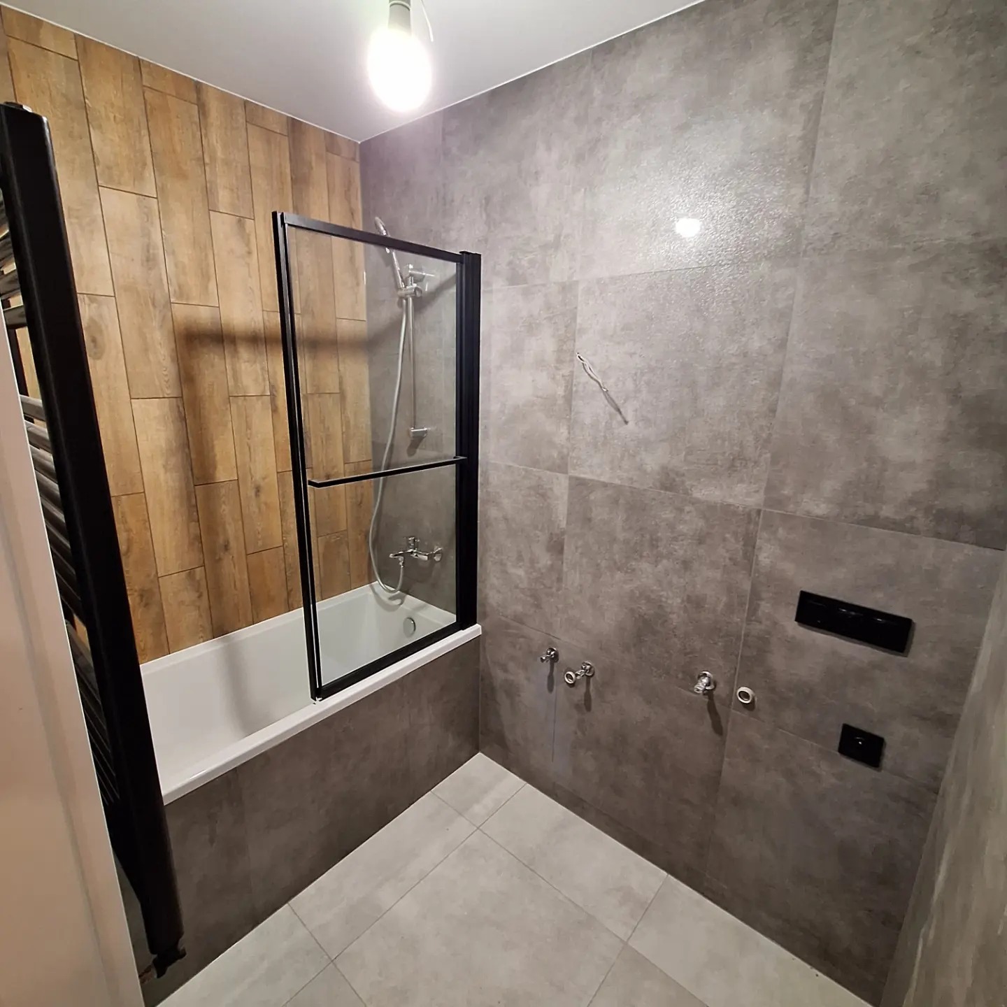 Nowoczesna łazienka z wanną obudowaną szarą płytką, przeszklonym parawanem nawannowym z czarną ramą, drewnopodobnymi płytkami na jednej ścianie oraz szarą podłogą i ścianami. Widoczne baterie...