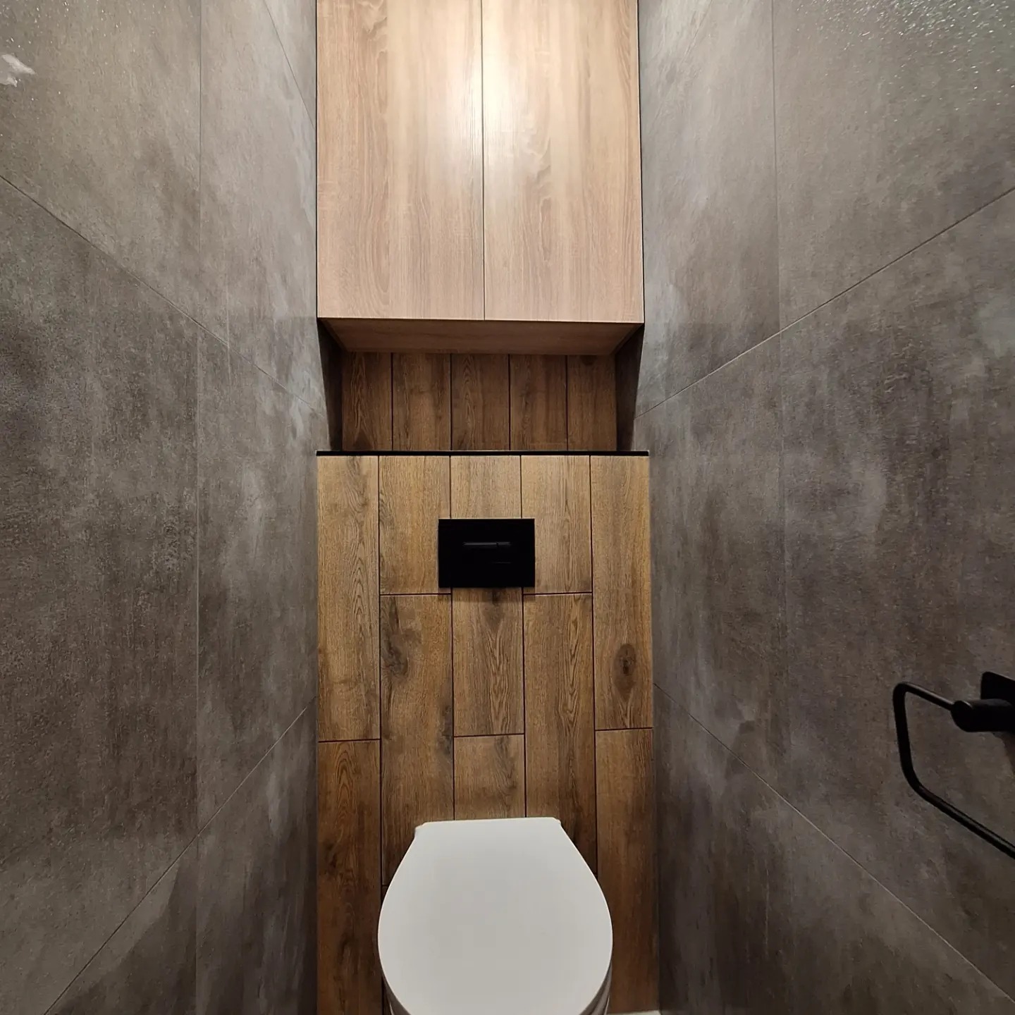 Nowoczesna toaleta z zabudową geberit wykończoną drewnopodobnymi panelami, szare płytki na ścianach i wisząca szafka nad stelażem.