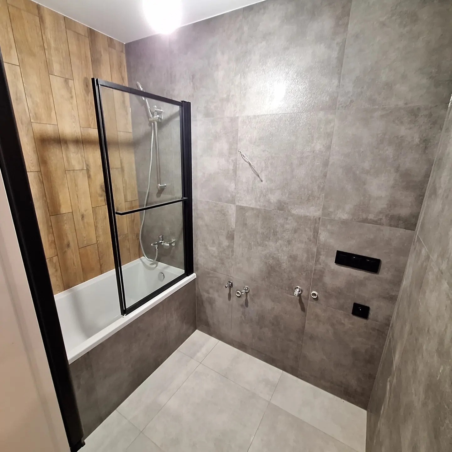 Wykończona łazienka z wanną obudowaną szarą płytką, przeszklonym parawanem nawannowym z czarną ramą, szarą płytką na ścianach i podłodze oraz drewnopodobnymi panelami na jednej ze ścian.
