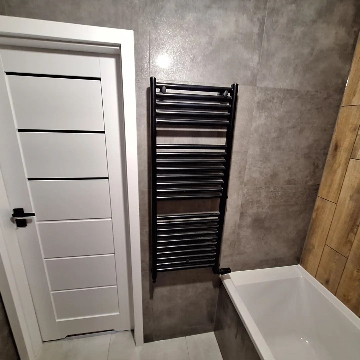 Nowoczesna łazienka z białą wanną, grzejnikiem drabinkowym w kolorze czarnym i białą, minimalistyczną, wewnętrzną stolarką drzwiową z czarną klamką na tle szarych płytek.