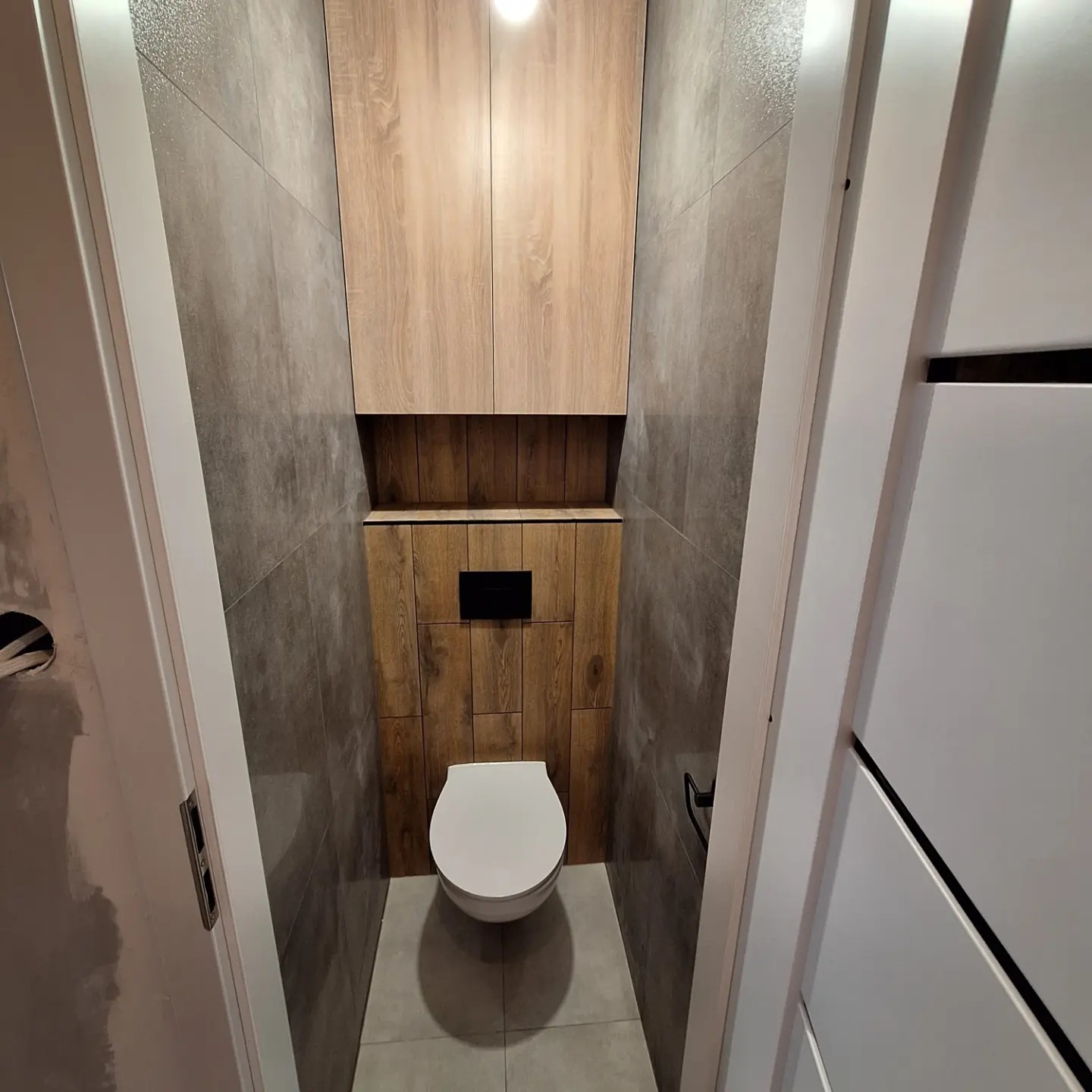 Wąska toaleta po remoncie z szarymi płytkami na ścianach, zabudową z drewnopodobnych paneli i szafką nad spłuczką, widok zza uchylonych białych drzwi.