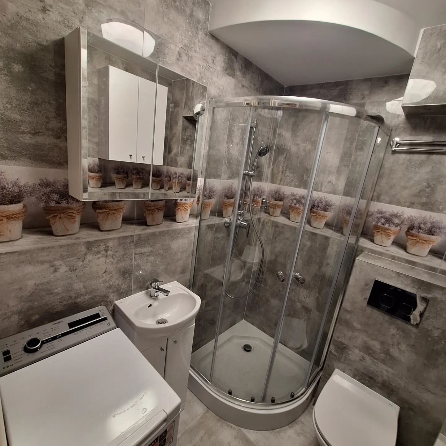 Narożna kabina prysznicowa z chromowanymi profilami w szarej łazience z dekoracyjnymi doniczkami na półce, obok umywalka z szafką i pralka.