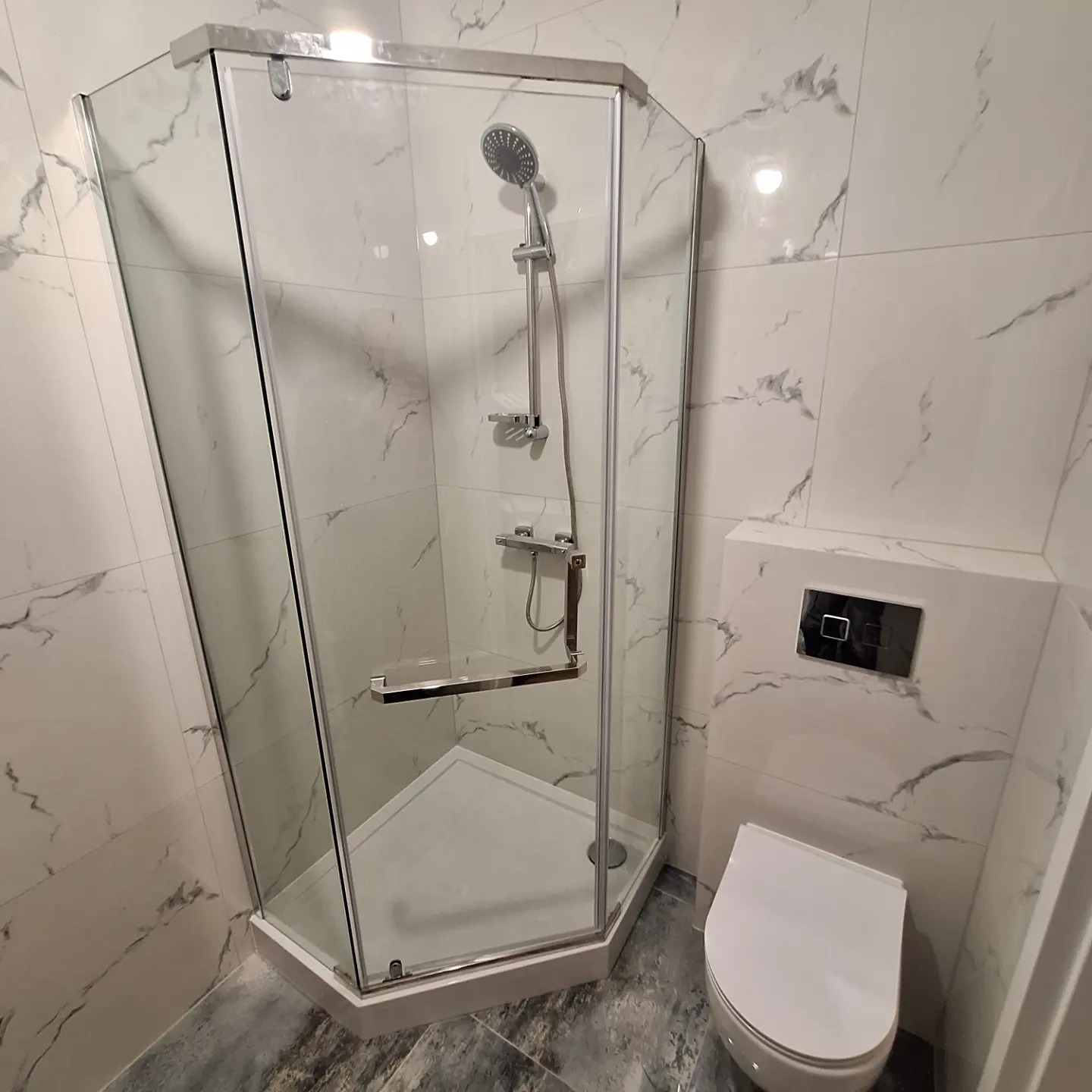 Nowoczesna kabina prysznicowa narożna z jasnymi płytkami imitującymi marmur, biała toaleta podwieszana i szara podłoga w łazience po remoncie.