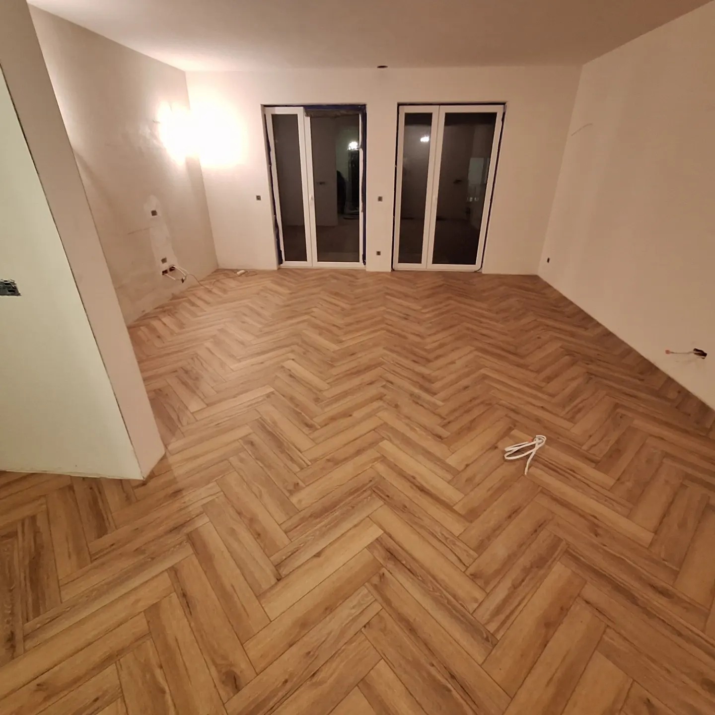 Nowo ułożona podłoga z paneli w jodełkę w pustym, jasnym pomieszczeniu z dwoma parami białych drzwi balkonowych; widoczne kable elektryczne wystające ze ściany.