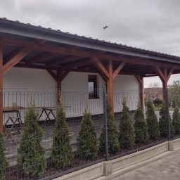 Centra - Zadaszona drewniana konstrukcja na betonowych słupach, widoczna zza metalowego ogrodzenia z tujami wzdłuż ścieżki z kostki brukowej, na tle białej ściany budynku i pochmurnego nieba z lecącym ptakiem.