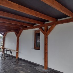 Centra - Zadaszenie tarasu z drewnianą konstrukcją nośną, widoczna biała ściana budynku z oknem i szara kostka brukowa na podłożu.