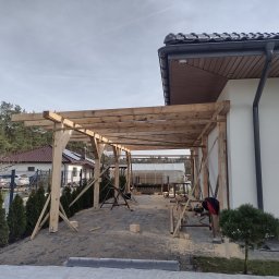 Centra - Drewniana konstrukcja zadaszenia tarasu w trakcie budowy, widoczne podpory i elementy mocujące, otoczenie w trakcie prac budowlanych.