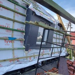 Centra - Montaż izolacji termicznej i folii paroizolacyjnej na dachu z widocznym oknem dachowym i stosem dachówek ceramicznych na budowie.
