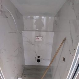 Centra - Łazienka w trakcie wykończenia, białe płytki imitujące marmur na ścianach, widoczne instalacje podtynkowe, na podłodze szary beton, oparta o ścianę drewniana poziomica.