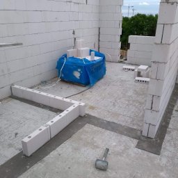 Centra - Wznoszenie ścian domu z bloczków z betonu komórkowego, widoczny fragment fundamentu i paleta z materiałem budowlanym zabezpieczona niebieską folią.