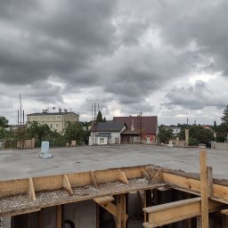Centra - Wylany strop betonowy z drewnianym szalunkiem w trakcie budowy domu murowanego, widok na okoliczne budynki i pochmurne niebo.