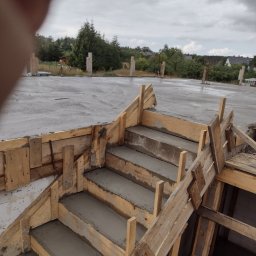Centra - Betonowe schody w drewnianym szalunku, świeżo wylane, na tle zalanego betonem fundamentu domu w budowie.