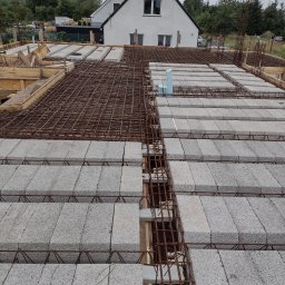 Centra - Wieniec stropowy w trakcie budowy, zbrojenie stalowe i betonowe bloczki szalunkowe na drewnianym szalunku, w tle dom jednorodzinny i pochmurne niebo.