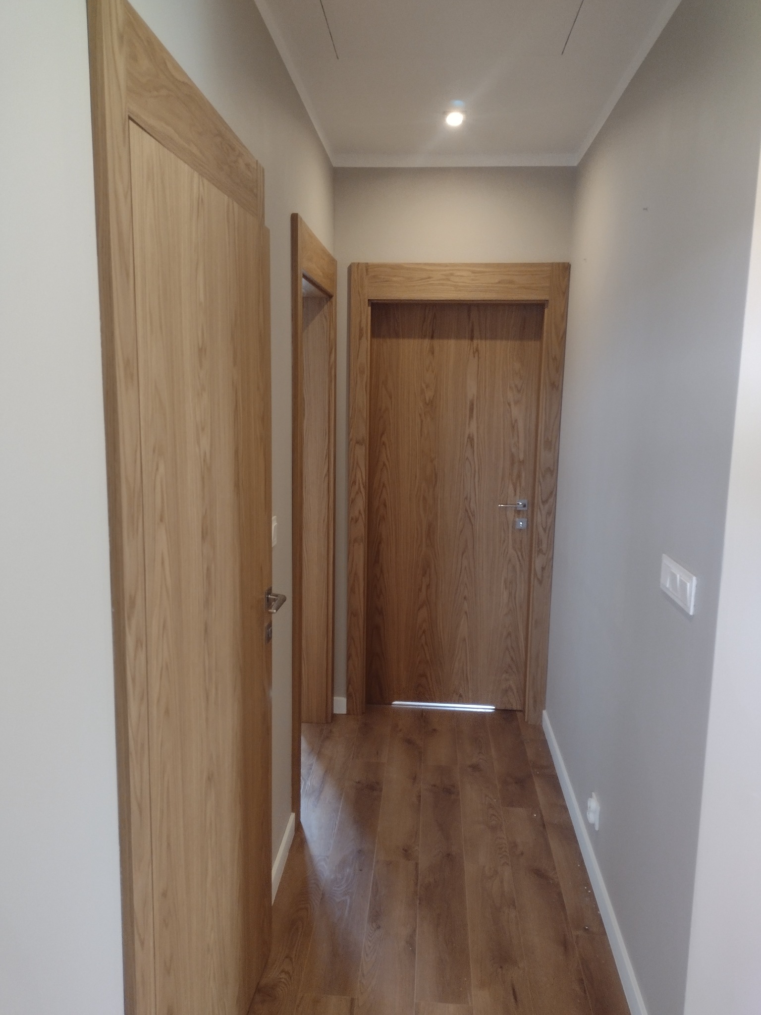 Wnętrze korytarza z trzema drewnianymi drzwiami o prostych ościeżnicach i panelami podłogowymi imitującymi drewno.