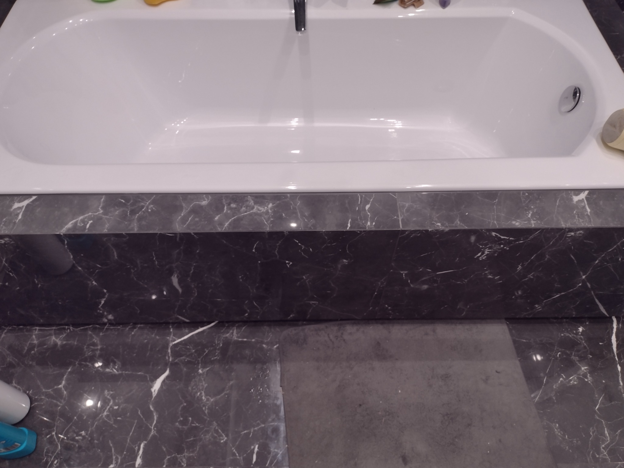 Biała wanna z chromowaną armaturą, obudowana czarnym marmurem z białymi żyłami, widoczne fragmenty podłogi z tego samego kamienia.