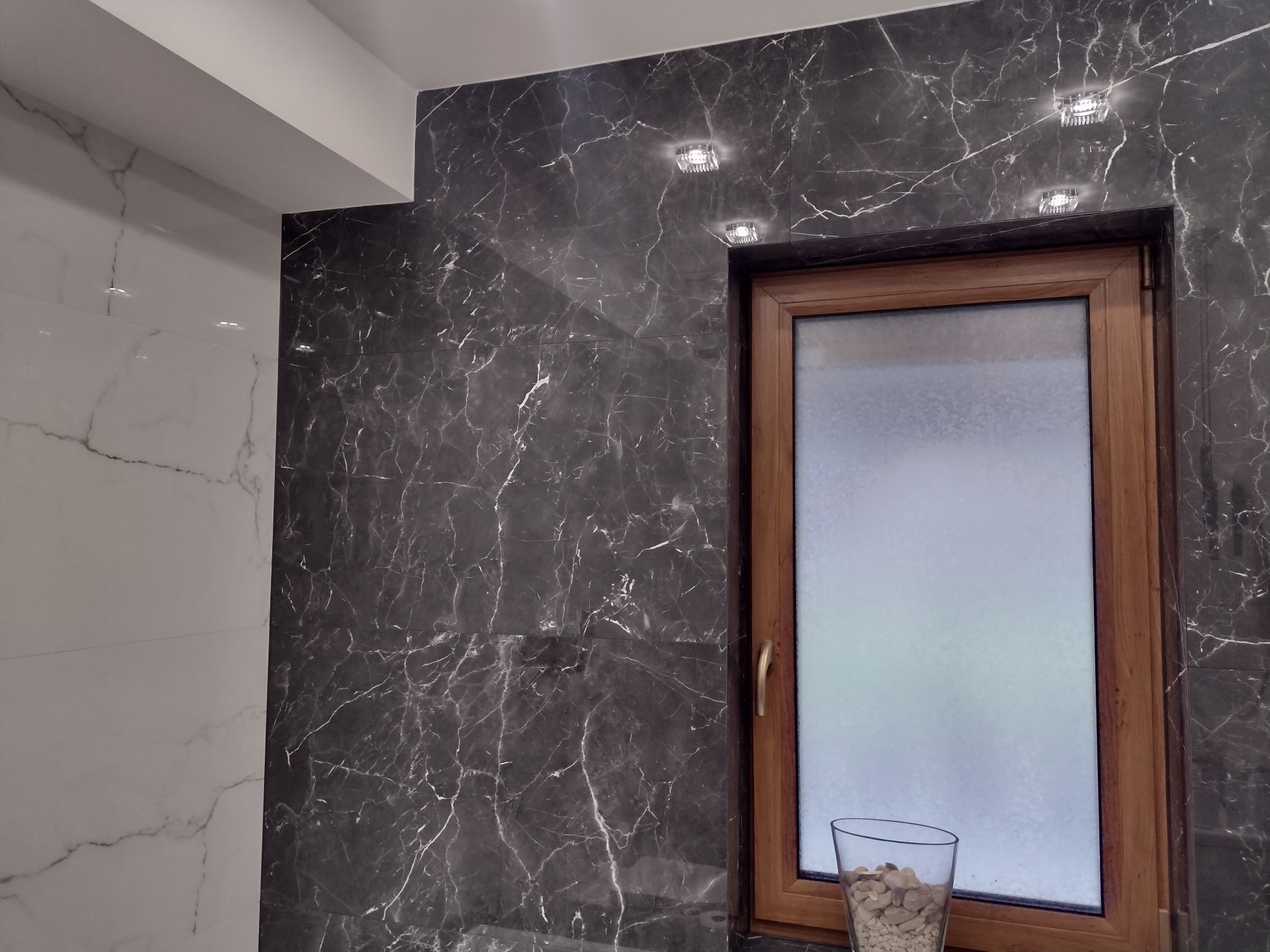 Wykończona łazienka z szarymi marmurowymi płytkami, drewnianą ramą okienną i szklanką z dekoracyjnymi kamieniami na parapecie.