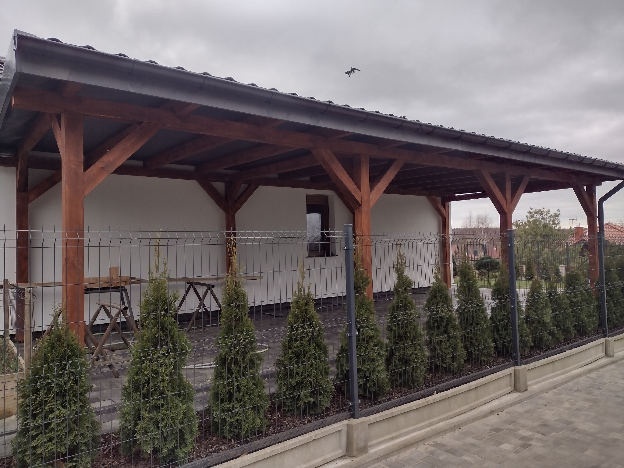 Zadaszona drewniana konstrukcja na betonowych słupach, widoczna zza metalowego ogrodzenia z tujami wzdłuż ścieżki z kostki brukowej, na tle białej ściany budynku i pochmurnego nieba z lecącym ptakiem.