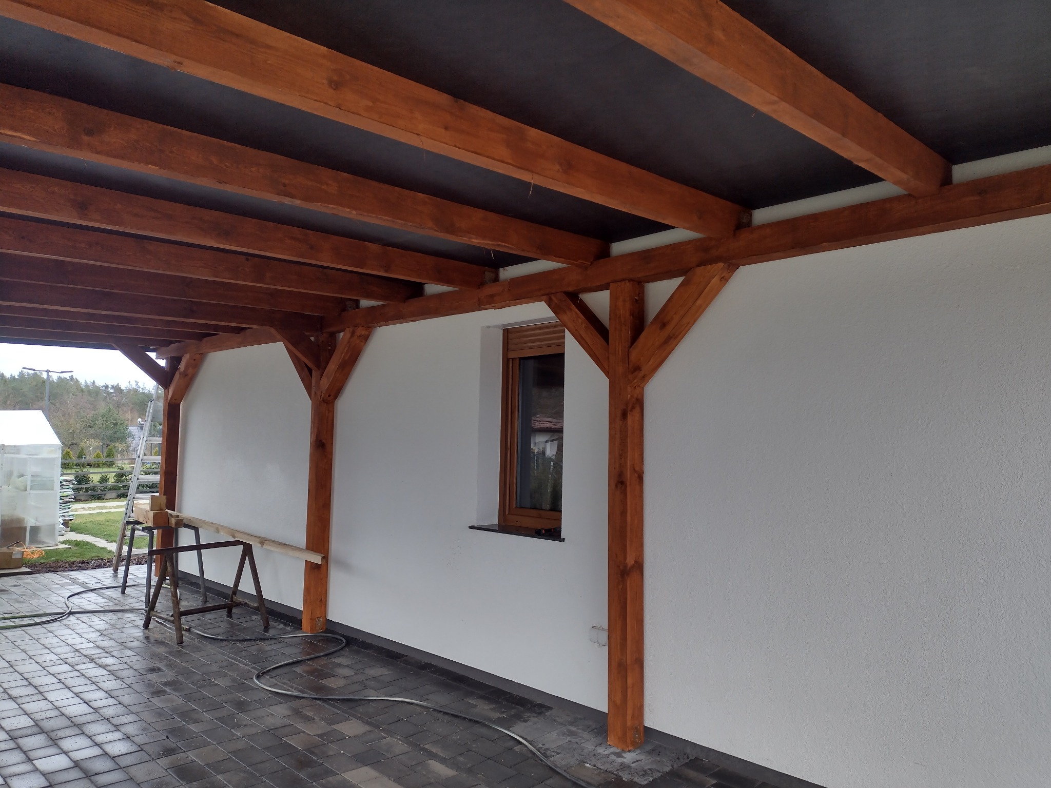 Zadaszenie tarasu z drewnianą konstrukcją nośną, widoczna biała ściana budynku z oknem i szara kostka brukowa na podłożu.