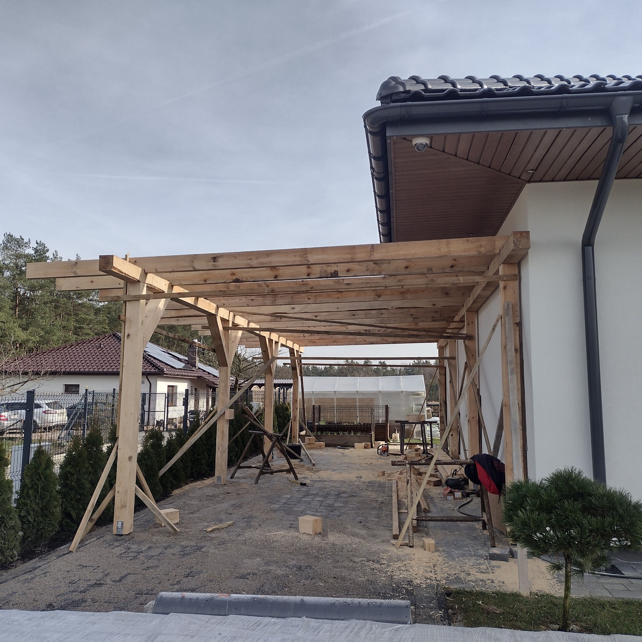 Drewniana konstrukcja zadaszenia tarasu w trakcie budowy, widoczne podpory i elementy mocujące, otoczenie w trakcie prac budowlanych.