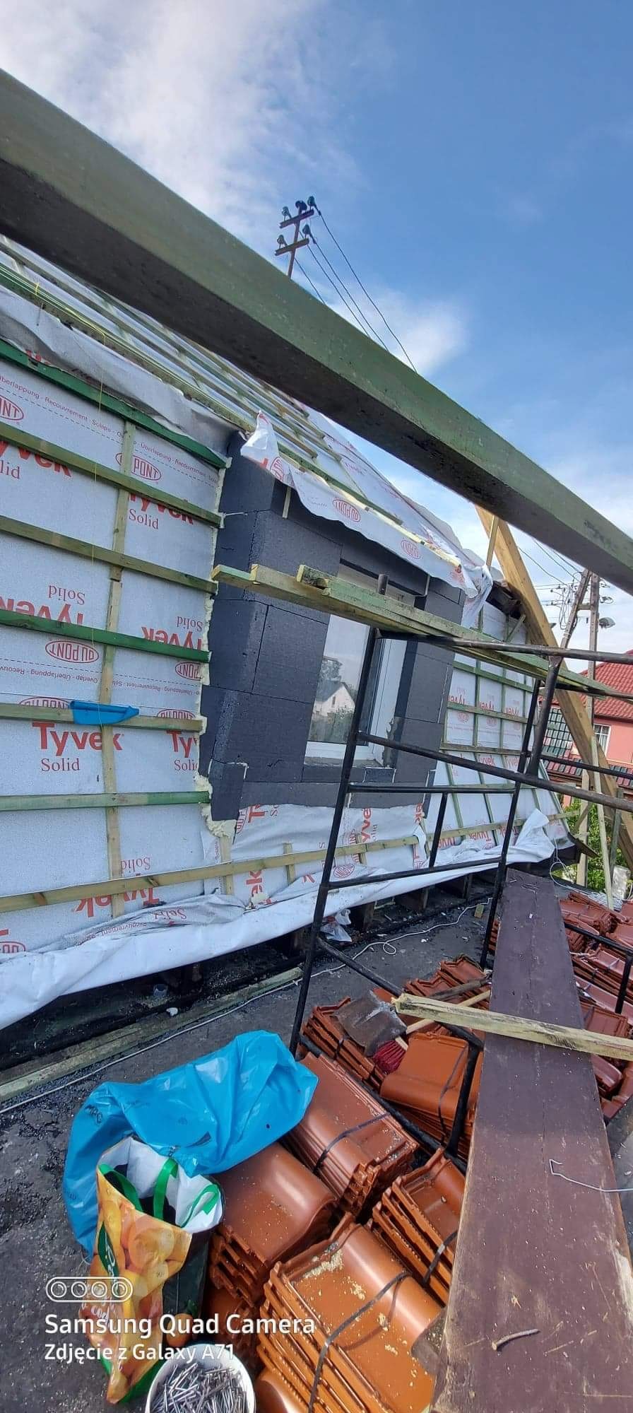 Montaż izolacji termicznej i folii paroizolacyjnej na dachu z widocznym oknem dachowym i stosem dachówek ceramicznych na budowie.