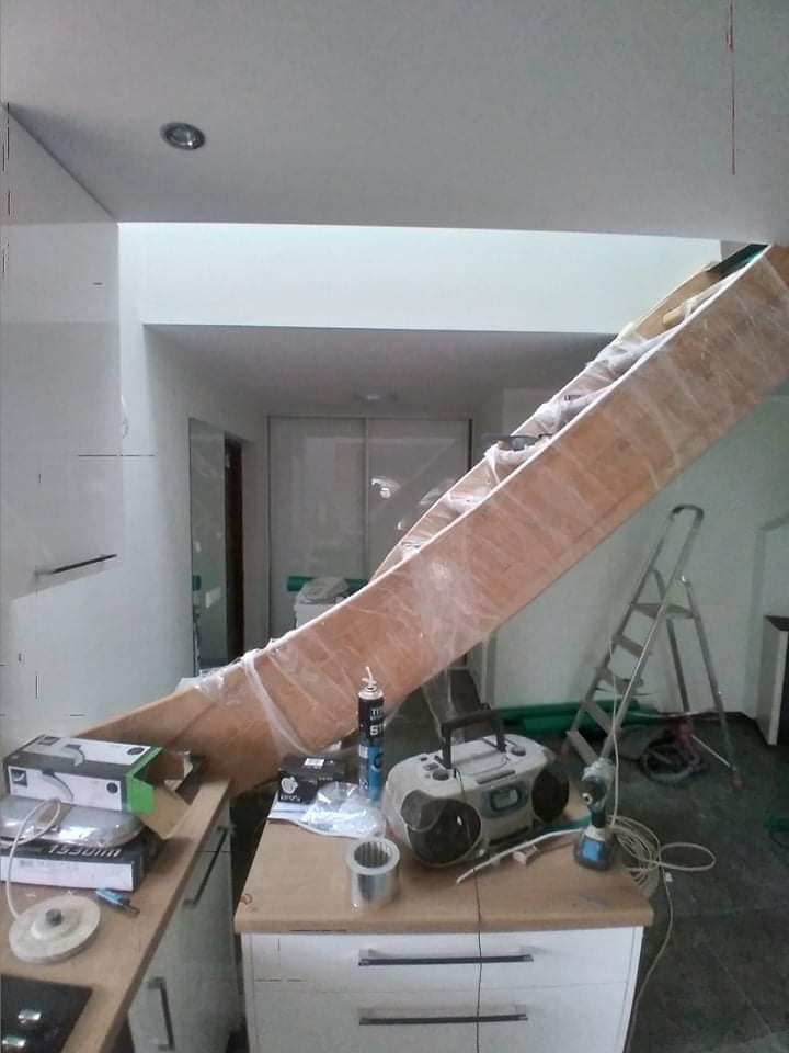 Wnętrze w trakcie remontu: zabezpieczone folią drewniane schody tymczasowo oparte o blat kuchenny, obok radio, puszka pianki montażowej i narzędzia, w tle drabina i białe ściany.