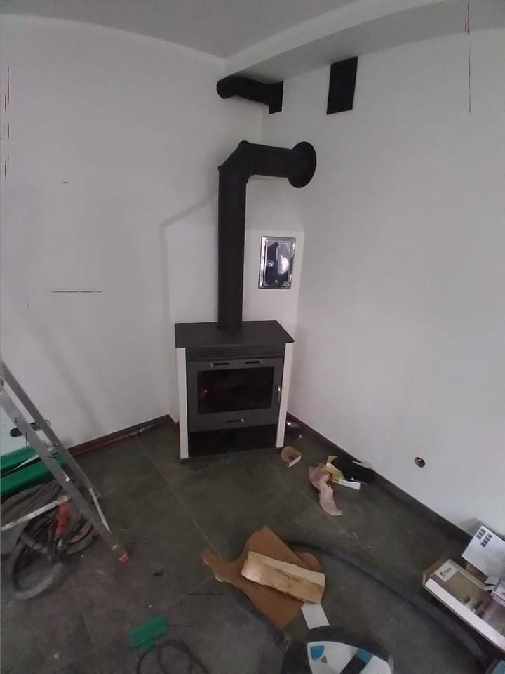 Montaż pieca wolnostojącego z czarnym kominem w rogu pomieszczenia, widoczne narzędzia i drabina.