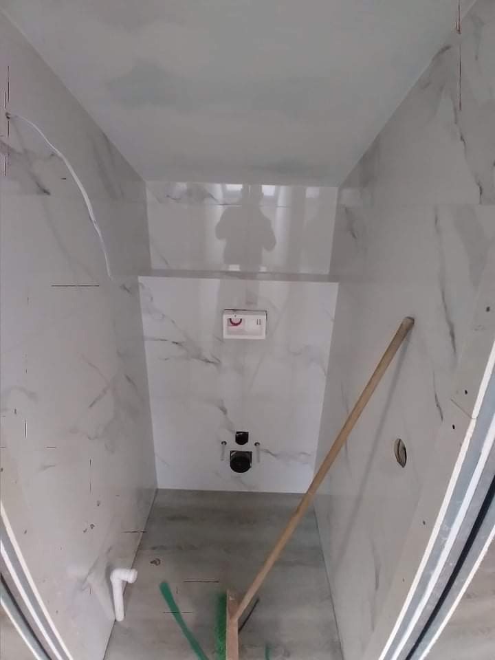 Łazienka w trakcie wykończenia, białe płytki imitujące marmur na ścianach, widoczne instalacje podtynkowe, na podłodze szary beton, oparta o ścianę drewniana poziomica.