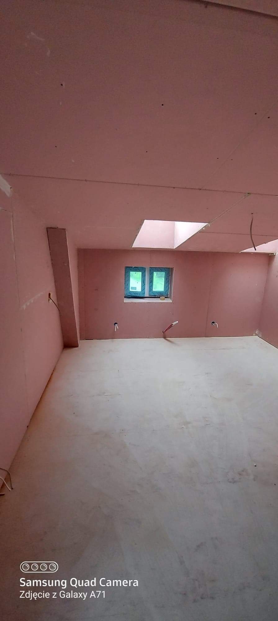 Surowe wnętrze pokoju z oknem i świetlikiem, ściany i sufit pokryte płytami kartonowo-gipsowymi, podłoga wylewka betonowa, widoczne przewody instalacji elektrycznej.