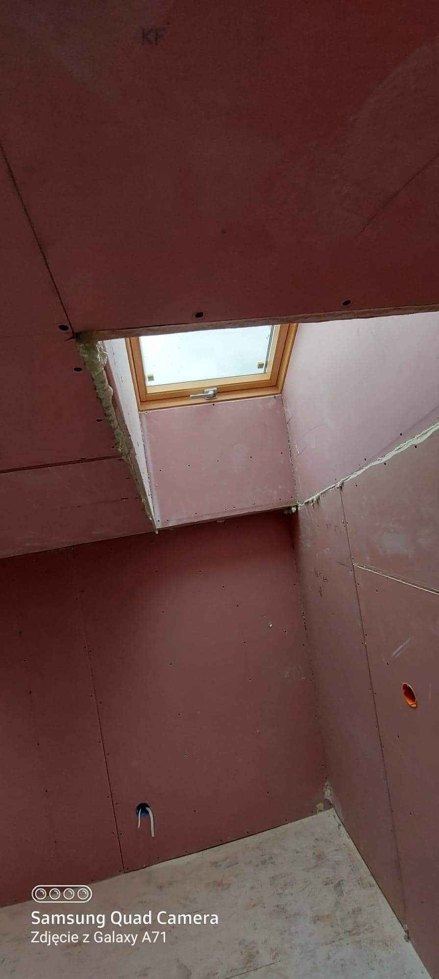 Wnętrze poddasza z zamontowanym oknem dachowym, płyty kartonowo-gipsowe na ścianach i suficie, widoczne ślady pianki montażowej wokół okna.