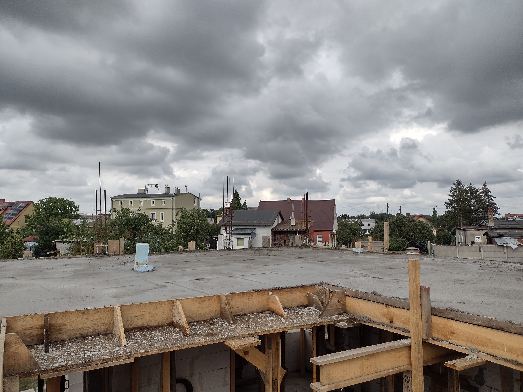 Wylany strop betonowy z drewnianym szalunkiem w trakcie budowy domu murowanego, widok na okoliczne budynki i pochmurne niebo.