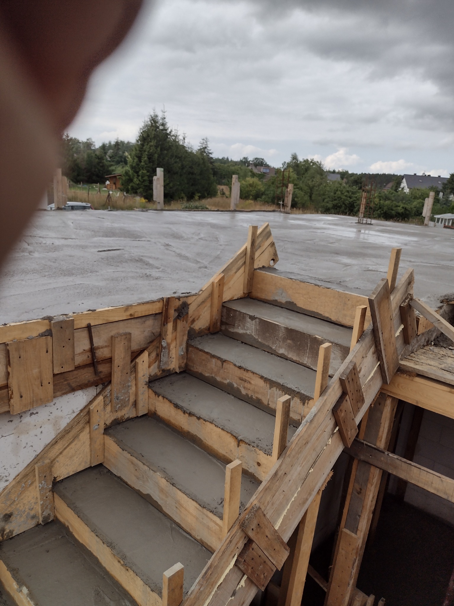 Betonowe schody w drewnianym szalunku, świeżo wylane, na tle zalanego betonem fundamentu domu w budowie.