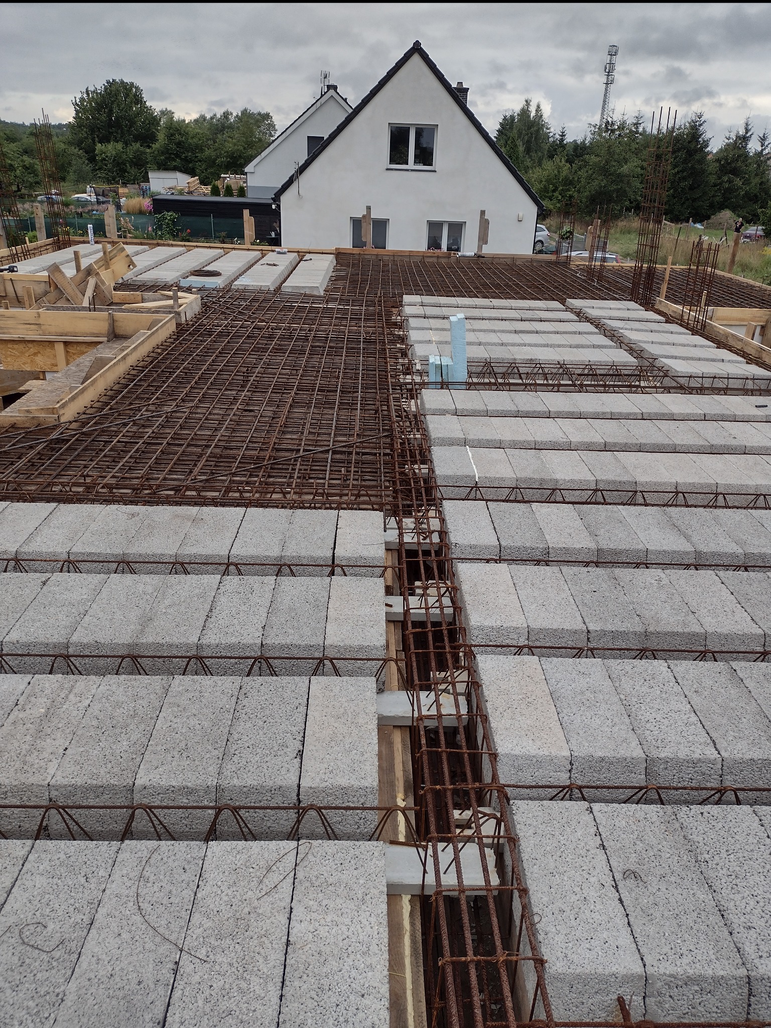 Wieniec stropowy w trakcie budowy, zbrojenie stalowe i betonowe bloczki szalunkowe na drewnianym szalunku, w tle dom jednorodzinny i pochmurne niebo.