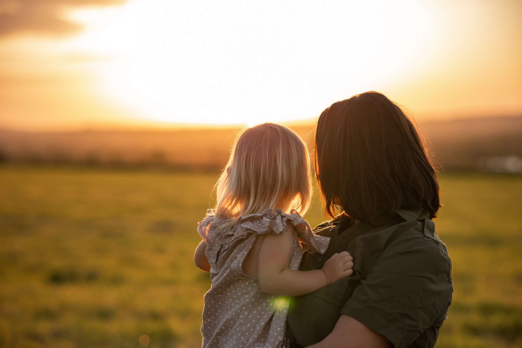 Matka trzymająca małą córeczkę na tle zachodzącego słońca nad polami, widok z tyłu, ciepłe oświetlenie.