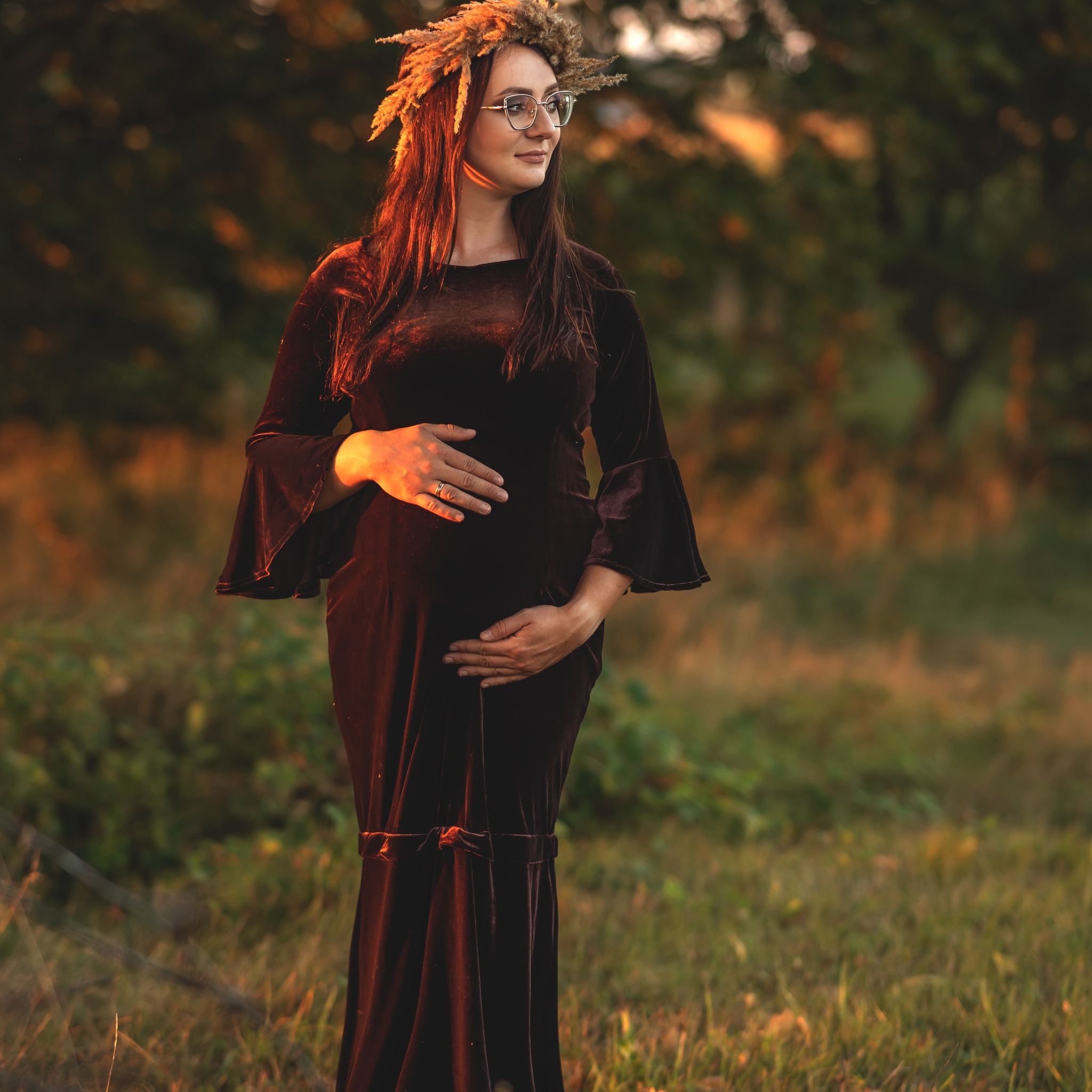 Kobieta w ciąży w aksamitnej sukni z wiankiem z trawy na głowie, delikatnie obejmuje brzuch na tle rozmytej zieleni i zachodzącego słońca.