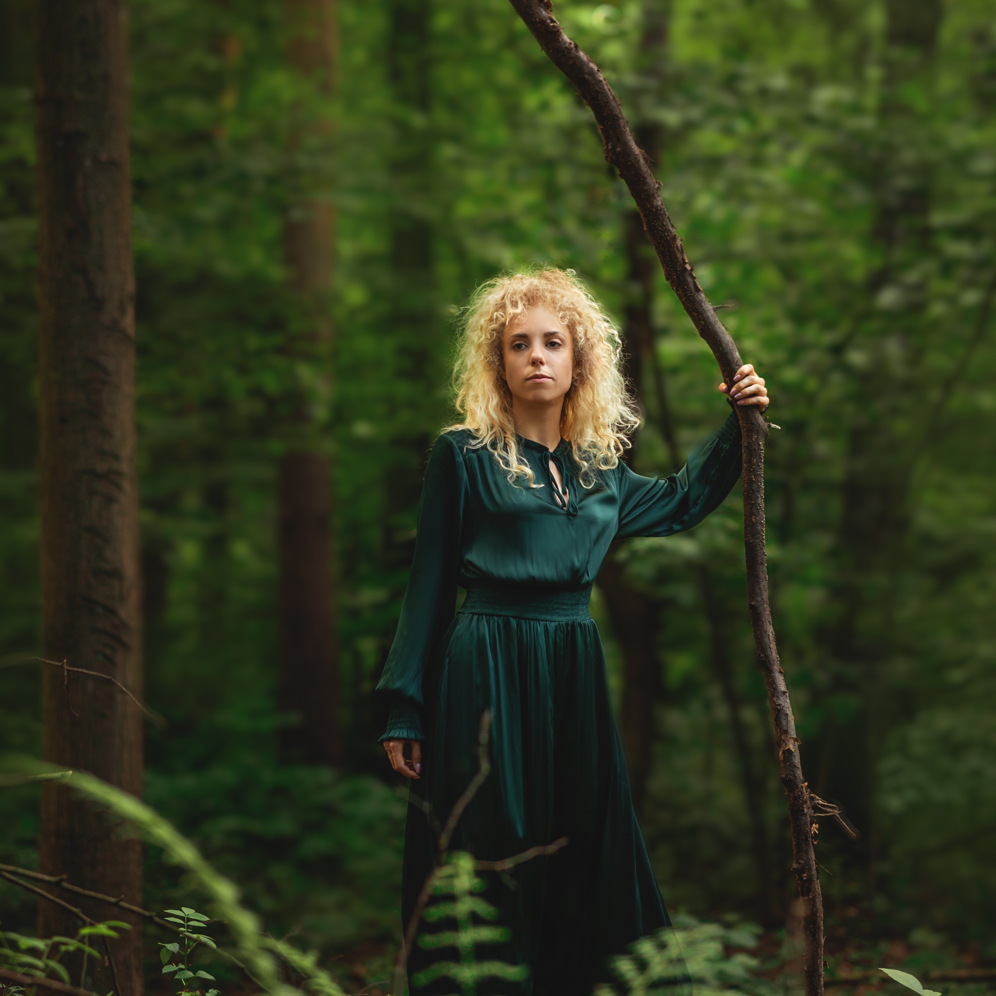 Portret kobiety o blond włosach w leśnej scenerii, ubrana w długą, ciemnozieloną suknię, opierająca się o gałąź drzewa.