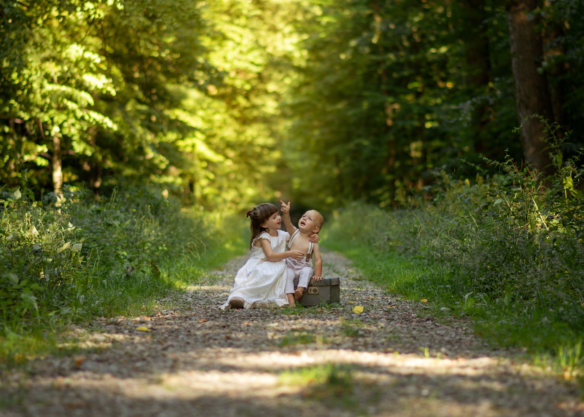 Dziewczynka w białej sukience siedzi obok niemowlęcia na starej walizce na leśnej ścieżce, wskazując palcem w górę, w tle rozmyte drzewa i zielona roślinność.