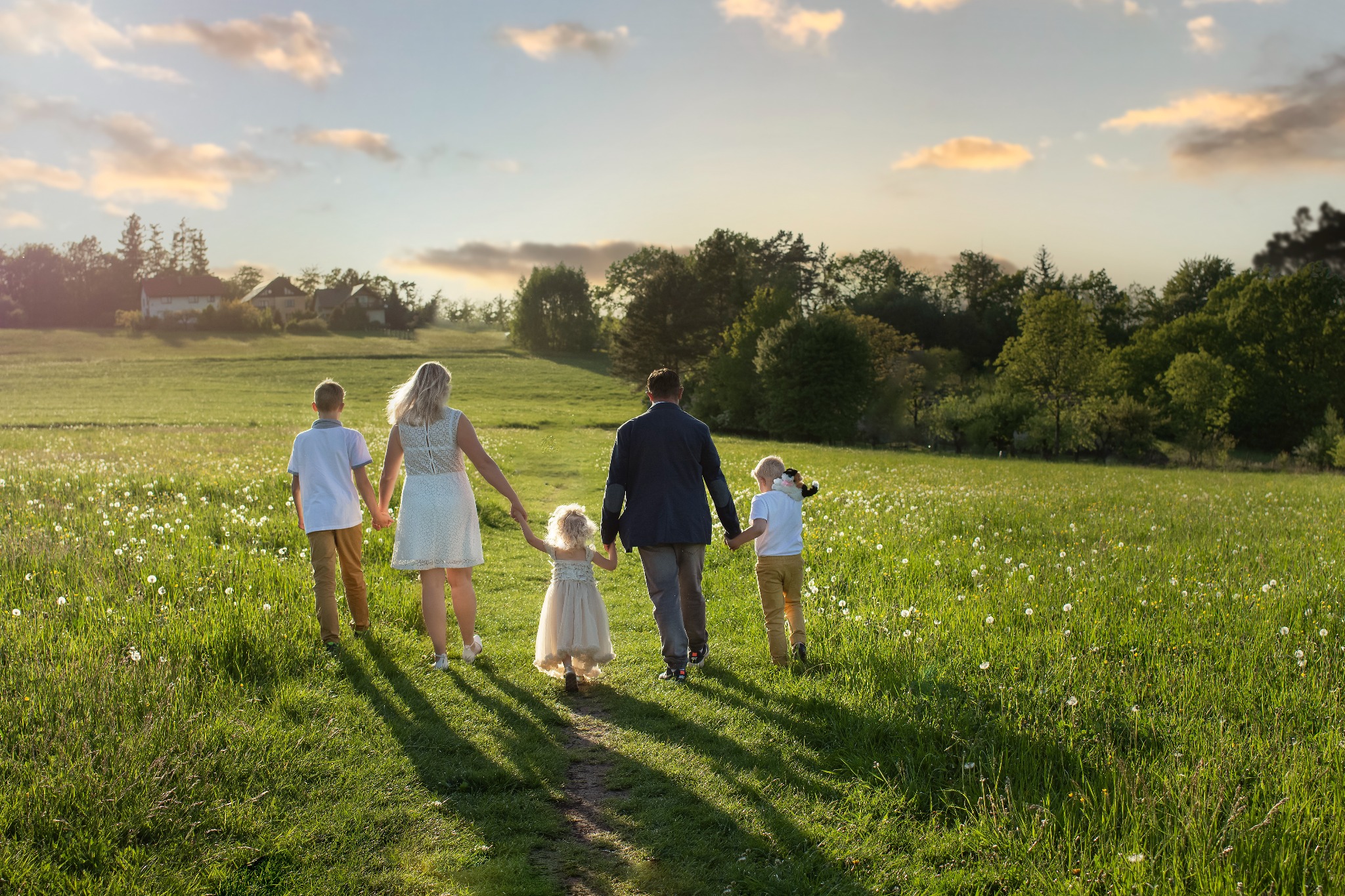 Rodzina z trojgiem dzieci idzie ścieżką przez łąkę w kierunku zachodzącego słońca, trzymając się za ręce; najmłodsza dziewczynka w tiulowej sukience.