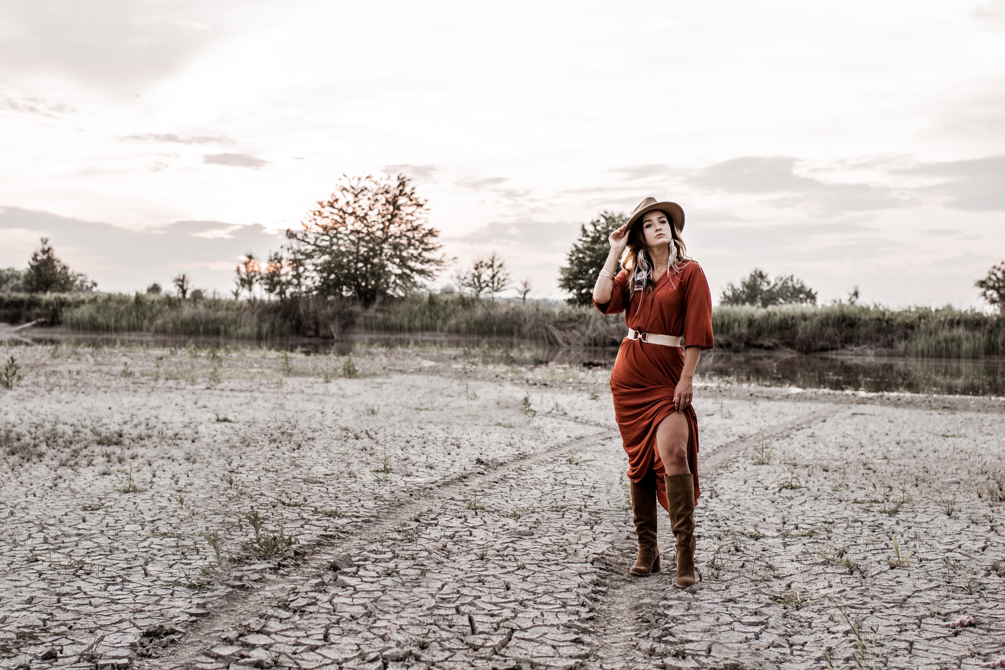 Kobieta w brązowej sukience i kapeluszu pozuje na spękanej, wyschniętej ziemi z rzadką roślinnością i drzewami w tle.