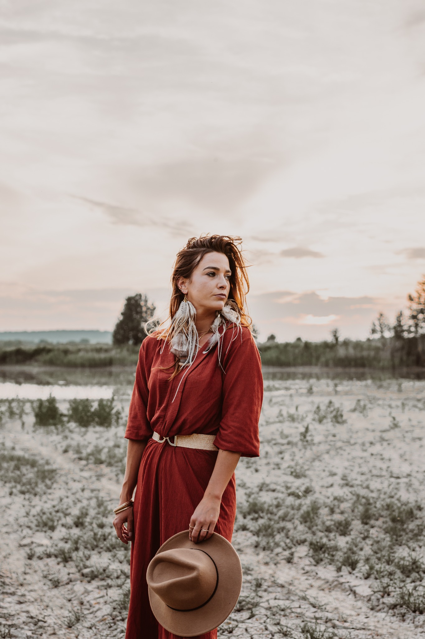 Portret kobiety w Ustroniu w długiej, ceglanej sukience, z kapeluszem w ręku i piórowymi kolczykami, na tle krajobrazu z jeziorem o zachodzie słońca.